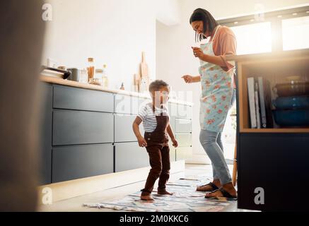 Ein kleiner gemischter Rennjunge, der tanzt und Spaß mit seiner Mutter in einer Küche hat. Glückliche Familie von zwei Kindern, die beim Backen in einer Küche Begeisterung ausdrückt Stockfoto