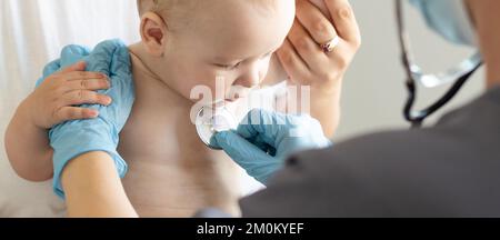 Ärztin untersucht kleines lächelndes Mädchen, das von der Mutter gehalten wird Stockfoto