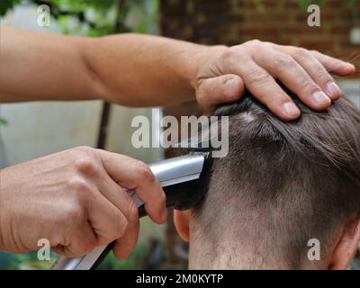 Ein Mann schneidet einem Teenager die Haare, schneidet lange dunkle Haare mit einem Trimmer ab, frisiert sie auf einer Plastikdüse, schneidet zu Hause mit einem Haarschneider Stockfoto