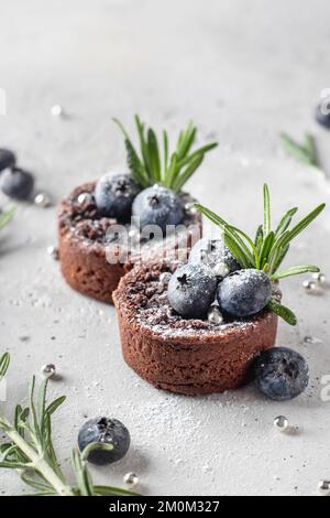 Zwei Schokoladenbrownies mit Blaubeeren und Rosmarin auf weißem, strukturiertem Hintergrund. Rezepte für Weihnachten und Neujahr. Vertikale Ausrichtung Stockfoto