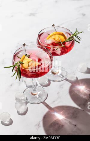 Zwei Weingläser Granatapfel, Eiscocktail mit Rosmarin und Zitrone auf weißem Marmorhintergrund, isoliert mit klaren Schatten in rauem Licht. Xm Stockfoto