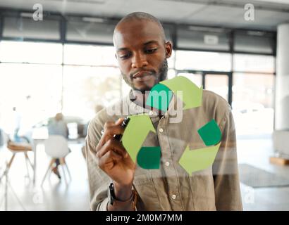 Junger fokussierter afroamerikanischer Geschäftsmann, der ein Recycling-Symbol auf ein Glasfenster in einem Büro zeichnet. Ein seriöser Geschäftsmann, der einen entwirft Stockfoto