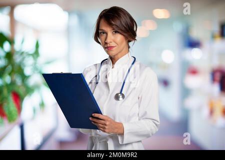 Eine Ärztin mittleren Alters, die einen weißen Laborkittel trägt, während sie in einem Krankenhausgebäude steht. Sie sieht in die Kamera und lächelt. Attraktive fema Stockfoto