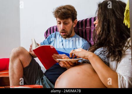 Nahaufnahme eines entspannten, bärtigen, weißen Erwachsenen, der seiner schwangeren Frau eine Geschichte vorliest, während sie Nüsse auf einem Teller isst und auf dem Sofa im l sitzt Stockfoto