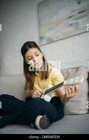 Brünettes Mädchen, das die kleine hawaiianische Gitarre singt, während es zu Hause auf dem Sofa sitzt Stockfoto