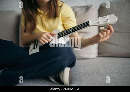 Kurzer Blick auf die verschwommene Ukulele des Kindes, während Sie auf dem Sofa mit gekreuzten Beinen sitzen Stockfoto