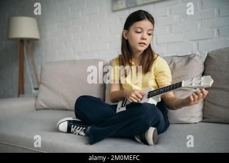 Braunes Mädchen, das auf der Couch sitzt, mit gekreuzten Beinen und einer kleinen hawaiianischen Gitarre Stockfoto