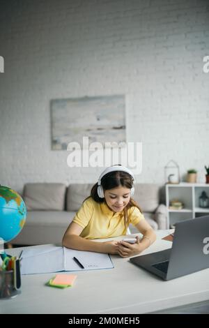 Positives Mädchen in Kopfhörern sitzt mit Smartphone in der Nähe des Laptops und Copybook auf dem Tisch im Wohnzimmer Stockfoto