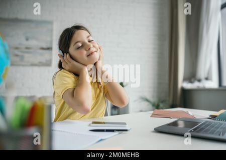 Zufriedenes Mädchen mit geschlossenen Augen, das Musik in Kopfhörern neben Notebook und Laptop auf verschwommenem Vordergrund hört Stockfoto