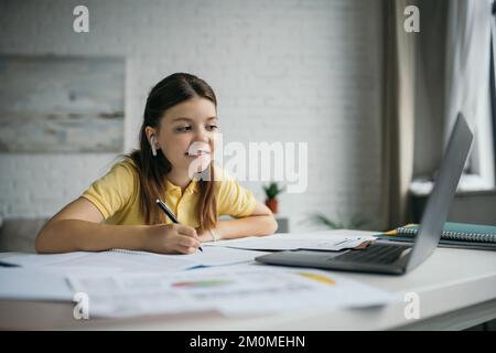 Lächelndes Mädchen in kabellosen Kopfhörern, das Stift hält und zu Hause auf ein Notebook schaut Stockfoto