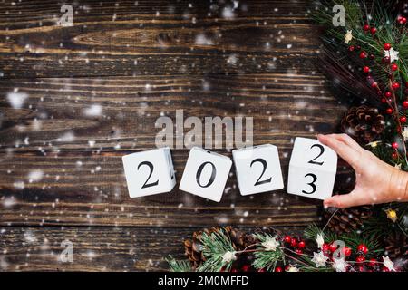 Frauenhand, die Silvester-Kalenderblöcke aus Holz für 2021 auf 2023 umwandelt. Weihnachtsbaumbeleuchtung, Kiefernäste, rote Winterbeeren und Schnee. Draufsicht. Stockfoto