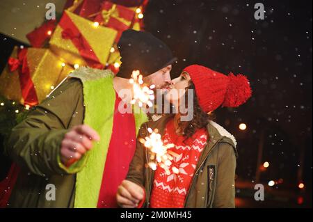 Ein Paar Liebhaber küsst sich vor dem Hintergrund von Weihnachtsgeschenken und Schneefällen und hält Funkeln in der Hand Stockfoto