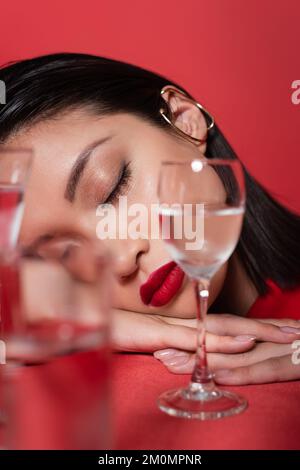 Porträt einer asiatischen Frau mit Make-up und geschlossenen Augen in der Nähe von Gläsern mit reinem Wasser auf verschwommenem Vordergrund isoliert auf rotem Stockbild