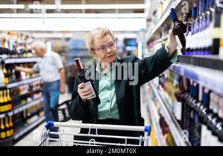 Reife Frau mit Gläsern wählt eine Flasche Wein in der Alkoholabteilung des Supermarkts Stockfoto