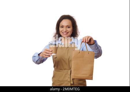 Die freundliche Barista hält ein Getränk zum Mitnehmen in einem Pappbecher bereit, lächelt beim Servieren der Bestellung für die Gäste nett und hat einen weißen Hintergrund Stockfoto