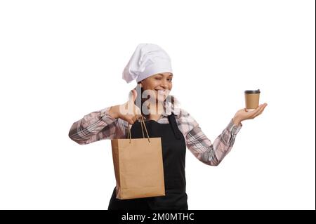 Die fröhliche Barista hält ein heißes Getränk zum Mitnehmen in einem Einwegbecher und einer recycelbaren Papiertüte bereit und zeigt den Daumen nach oben auf der Kamera Stockfoto