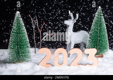 Kreative 2023-Karte. Ein glänzendes Silberhirsch, das nachts im Schnee im Wald zwischen den Weihnachtsbäumen auf schwarzem Hintergrund steht. Der Weihnachtsmann ist rei Stockfoto