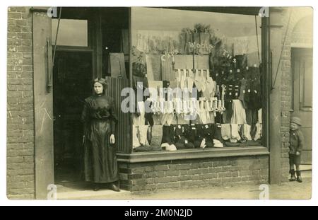 Originale charmante Postkarte aus der Zeit der edwardianischen und titanischen Ära von einer Eigentümerin oder Verkäuferin, die vor einem Herrenausstatter-Geschäft steht, in dessen Fenster der Verkauf von Krawatten, Hemden, Wollen und Strumpfwaren angekündigt wird. Das Kind eines Nachbarn blickt von einer Tür um die Ecke. Ein klassischer Moment in der Zeit. Edwardianischer Ladenbesitzer. Viktorianischer Ladenbesitzer. Die Postkarte stammt vom 31. Mai 1911 aus Warrington, Cheshire, England, Großbritannien Stockfoto