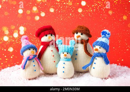 Eine Familie gestrickter Schneemänner feiert die Feiertage auf rotem Hintergrund mit goldenen Lichtern. Wintermärchen-Geschichte mit eigenen Händen, Hobby. Merry Chr Stockfoto