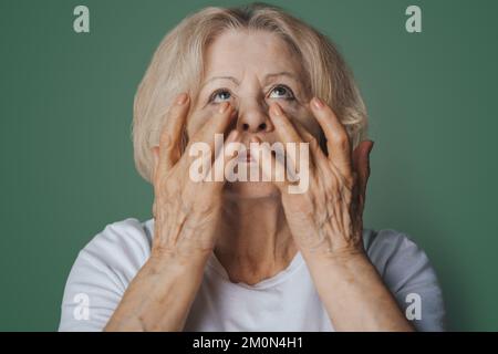 Porträt einer hübschen Seniorin mit Hydrogel-Unteraugenflecken am Morgen, mit einer Wirkung von perfekter Haut, isoliert auf grünem Hintergrund. Anti Stockfoto