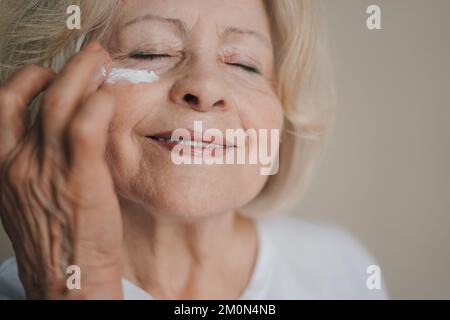 Glückliche, reife Frau, die Anti-Aging-Lotion aufträgt, um dunkle Kreise unter den Augen zu entfernen. Make-up-Produkt. Lächeln Sie. Stockfoto