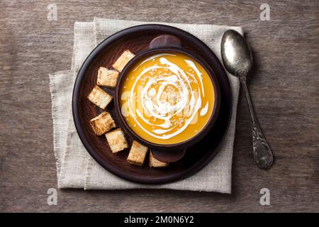 Kürbissuppe mit Sahne und Sesamsamen in brauner Keramikschüssel auf Holzhintergrund. Flach liegend, Draufsicht, Kopierbereich Stockfoto