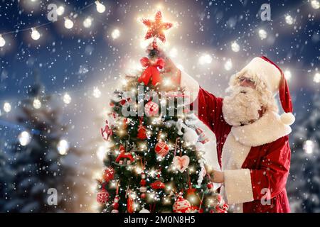 Der Weihnachtsmann schmückt den Weihnachtsbaum. Weihnachtsmann mit leuchtendem Stern auf dem dekorierten Weihnachtsbaum. Zauberhafte Winterferien. Geschenke und Geschenke. Stockfoto