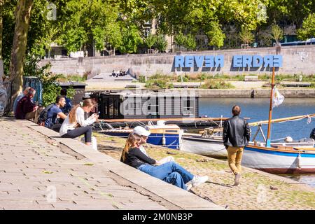 Junge Menschen genießen den Sonnenschein am Ufer des Flusses Erdre in Nantes, Frankreich, mit dem großen Schild „REVER ERDRE“ im Hintergrund. Stockfoto