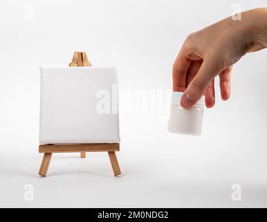 Leinwand-Modell auf winziger Staffelei aus Holz, Künstler Hand mit einem leeren Glas mit Gouache-Farbe. Minimalistische Kreativwerkzeuge, Zeichenmaterial. Hochwertiges Foto Stockfoto