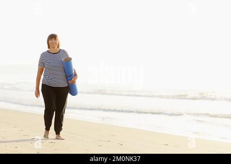 Frau im mittleren Alter, die eine Sportmatte hält und sich auf Yoga am Strand vorbereitet. Glückliche reife übergewichtige Frau, die am Meer trainiert Stockfoto