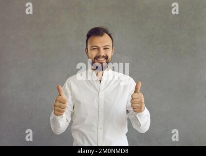 Porträt eines glücklichen jungen Mannes, der die Daumen nach oben hält, isoliert auf grauem Hintergrund Stockfoto