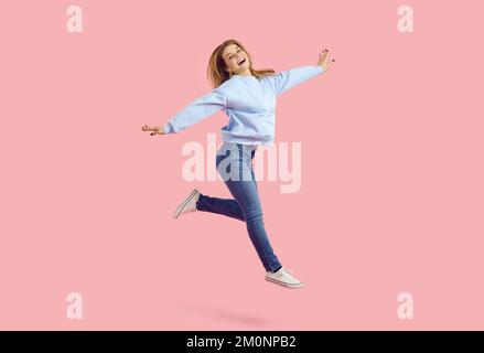 Lächelnde junge Frau, die im Studio mit ausgestreckten Armen auf pastellrosa Hintergrund Spaß hat. Stockfoto