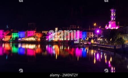 Porto Ceresio, Italien: 12-25-2021: Häuser beleuchtet durch Scheinwerfer in der Weihnachtsnacht, durch farbige Lichter, die auf das Wasser des Luga-Sees reflektieren Stockfoto