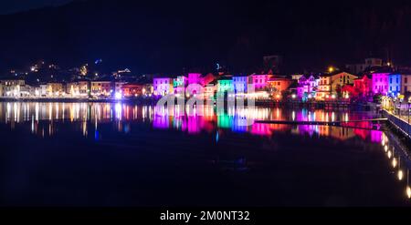 Porto Ceresio, Italien: 12-25-2021: Häuser beleuchtet durch Scheinwerfer in der Weihnachtsnacht, durch farbige Lichter, die auf das Wasser des Luga-Sees reflektieren Stockfoto