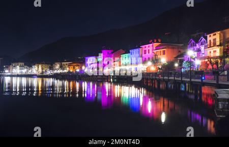 Porto Ceresio, Italien: 12-25-2021: Häuser beleuchtet durch Scheinwerfer in der Weihnachtsnacht, durch farbige Lichter, die auf das Wasser des Luga-Sees reflektieren Stockfoto