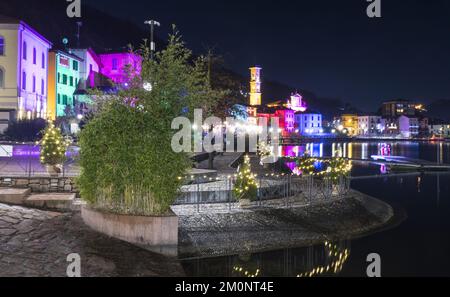 Porto Ceresio, Italien: 12-25-2021: Häuser beleuchtet durch Scheinwerfer in der Weihnachtsnacht, durch farbige Lichter, die auf das Wasser des Luga-Sees reflektieren Stockfoto