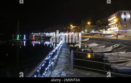 Portovaltravaglia, Italien - 12-21-2021: Kleiner Hafen, der von Weihnachtslichtern beleuchtet wird, die auf dem See reflektieren Stockfoto
