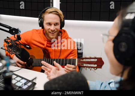 Ein junger weißer Mann spielt Gitarre, während eine glückliche Frau Podcast im Radio-Studio aufnimmt Stockfoto