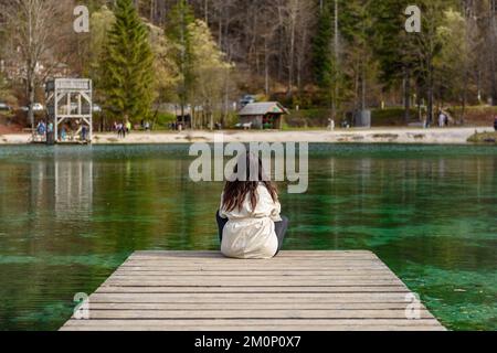 Ein Rückbild einer jungen Frau, die auf einem hölzernen Pier am Ufer des Jasna-Sees in Slowenien sitzt Stockfoto