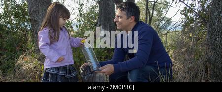 Bild eines lächelnden Vaters und einer lächelnden Tochter, während sie einen Hain von Müll und Plastikflaschen aufräumen. Umweltfreundliche Familie und Recycling. Stockfoto