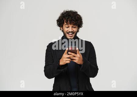 Ein junger, glücklicher, lächelnder indianer, der auf seinem Handy vor einem isolierten grauen Hintergrund zusieht Stockfoto