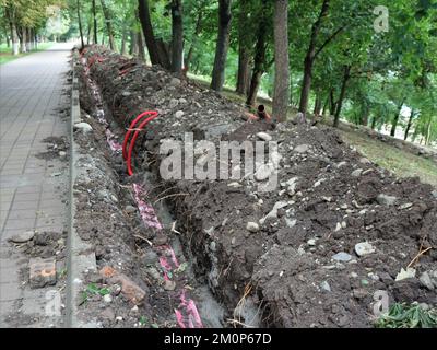 Arbeitet in einem Stadtpark und verlegt elektrische Kommunikation unterirdisch, ein Graben entlang eines gepflasterten Weges in einem Park mit roten Kunststoffrohren mit Drähten Stockfoto