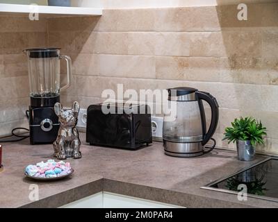 Elektrischer Wasserkocher, Toaster und Mixer auf der Theke in der Küche. Beige Fliesen Stockfoto