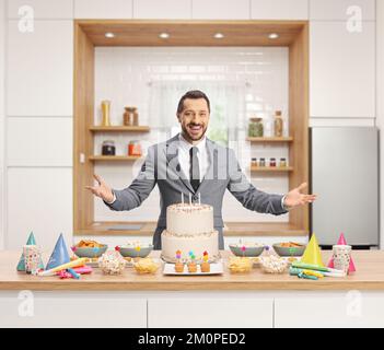 Junger Mann, der in einer Küche posiert, mit Kuchen und Partyzubehör auf einer Holztheke Stockfoto