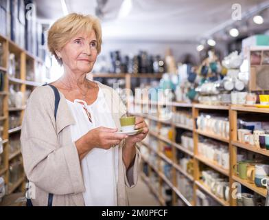 Reife weibliche Käufer wählt Tassen für Tee oder Kaffee im Geschirrladen Stockfoto