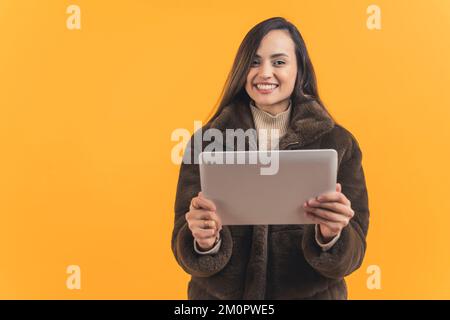 Fröhliche lateinamerikanische Frau in einem Wintermantel mit Tablet, orangefarbener Hintergrund, Mittleres Shot-Technologiekonzept. Hochwertiges Foto Stockfoto