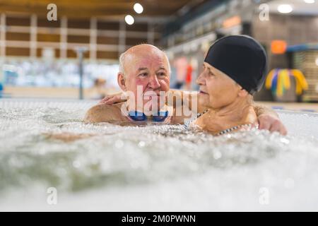 Älteres, weißes Ehepaar mit Schwimmbadausstattung - Haarschutzkappe und Brille - umarmte und unterhielt sich, während man in einem heißen Wasser saß. Hochwertiges Foto Stockfoto