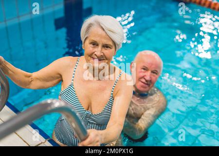 Eine reizende, positive, weiße Seniorin in gestreiftem Badeanzug kommt aus dem Pool. Kaukasischer Glatzkopf tauchte im Hintergrund in Wasser. Hochwertiges Foto Stockfoto