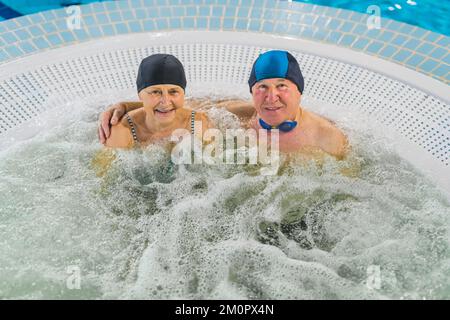 Ein bezauberndes, glückliches, weißes, verheiratetes Seniorenpaar, das sich im Whirlpool entspannen kann. Schwimmausstattung: Kappen, Gläser und Badebekleidung. Hochwertiges Foto Stockfoto