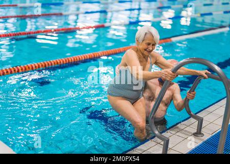 Lachende, positive, weiße Seniorin in gestreiftem Badeanzug, die versucht, mit der Unterstützung ihres unbekannten Mannes aus dem Pool zu kommen. Hochwertiges Foto Stockfoto